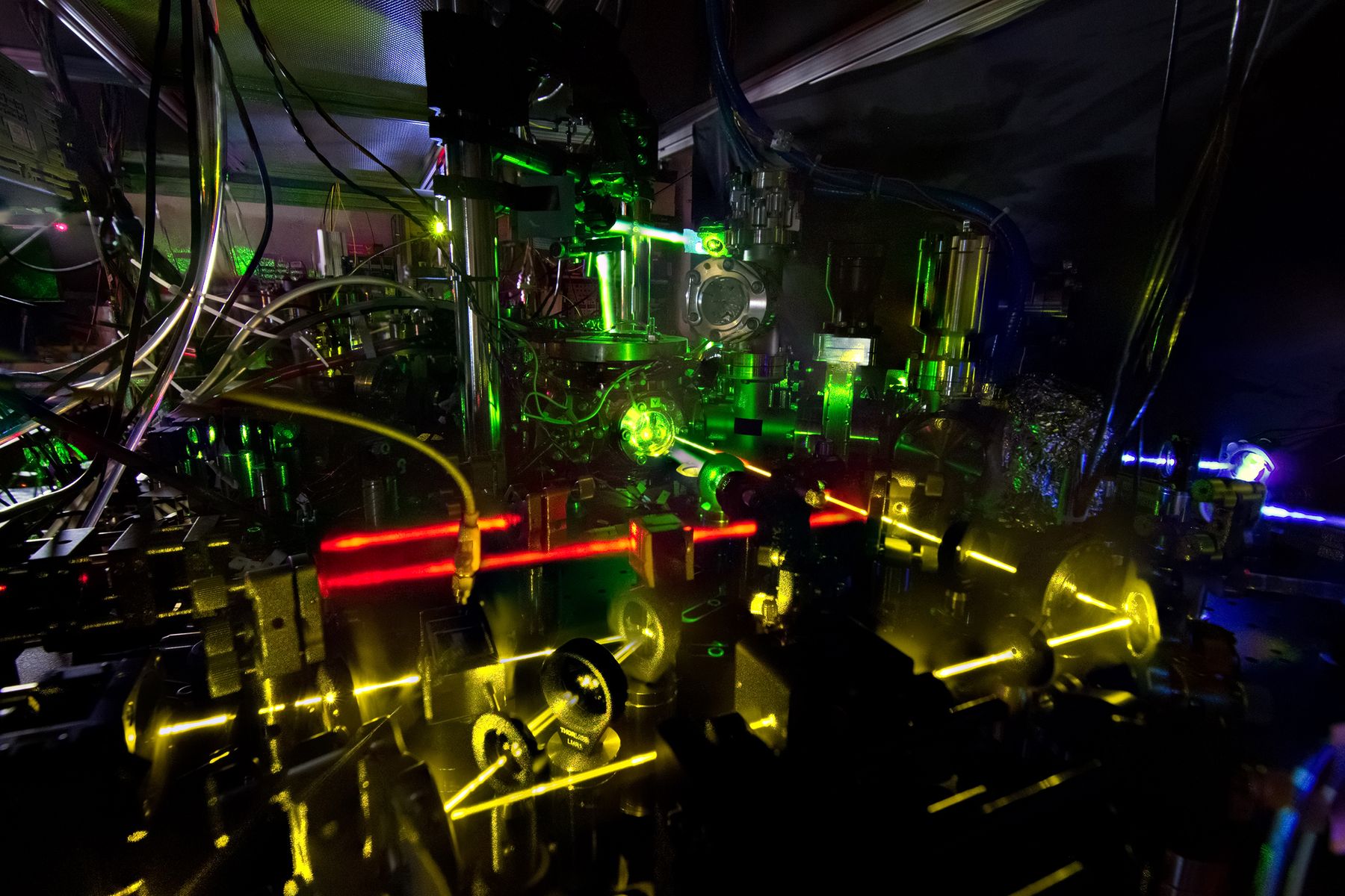 One of NIST's ytterbium lattice atomic clocks in a darkened laboratory: green, yellow, red and blue laser beams tracing paths between mirrors and vacuum hardware on the optical bench.